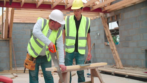 Carpenter-With-Male-Apprentice-Cutting-Wood-On-Building-Site