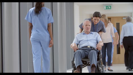 Enfermera-Empujando-Paciente-Senior-En-Silla-De-Ruedas-A-Lo-Largo-Del-Corredor