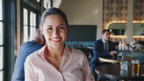 Portrait-Of-Female-Manager-Of-Busy-Cocktail-Bar-In-Restaurant-With-Customers-Smiling-At-Camera