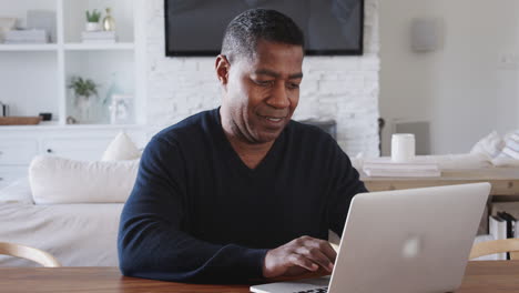Middle-aged-African-American-man-sitting-at-a-table-using-laptop-computer-at-home,-close-up,-zoom-out