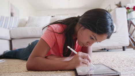 Teenager-Mädchen-Liegt-Im-Wohnzimmer-Auf-Dem-Boden-Und-Benutzt-Einen-Tablet-Computer-Und-Einen-Stift,-Fokus-Auf-Den-Vordergrund