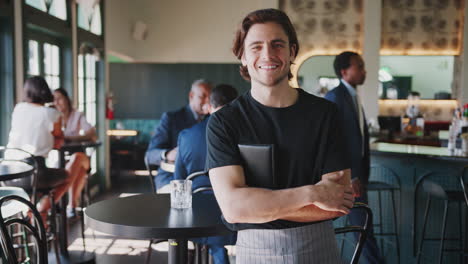 Portrait-Of-Waiter-In-Busy-Cocktail-Bar-Of-Restaurant-With-Customers-In-Background