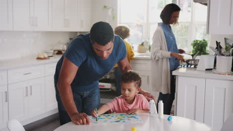 Dad-watches-son-painting-in-kitchen,-returns-to-chores-with-mother-and-daughter-in-the-background