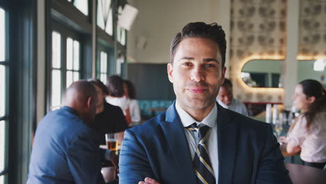 Portrait-Of-Smiling-Businessman-In-Busy-Cocktail-Bar-Of-Restaurant-With-Customers-In-Background