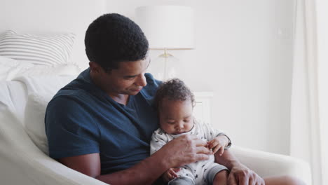 Millennial-African-American-dad-sitting-in-an-armchair-holding-his-baby-boy,-close-up,-side-view