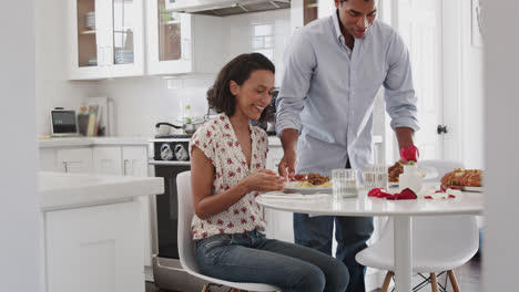 Junge-Erwachsene-Frau-Sitzt-Am-Tisch-In-Der-Küche,-Während-Ihr-Partner-Ein-Romantisches-Essen-Serviert