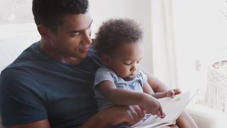 Side-view-of-millennial-African-American-father-sitting-and-reading-a-book-with-his-toddler-son,-close-up
