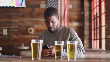 Hombre-Sentado-En-Un-Bar-Deportivo-Bebiendo-Cerveza-Y-Mirando-El-Teléfono-Móvil