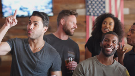 Group-Of-Male-And-Female-Friends-Celebrating-Whilst-Watching-Game-On-Screen-In-Sports-Bar