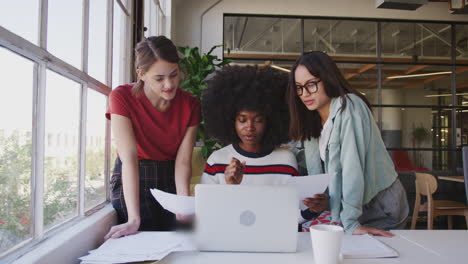 Three-millennial-women-working-together--with-a-laptop-at-a-desk-in-a-creative-office,-front-view