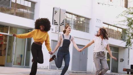 Drei-Millennial-Freundinnen-Halten-Sich-An-Den-Händen-Und-Lachen,-Während-Sie-über-Einen-Fußgängerüberweg-Laufen