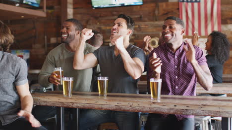 Group-Of-Male-Friends-Celebrating-Whilst-Watching-Game-On-Screen-In-Sports-Bar