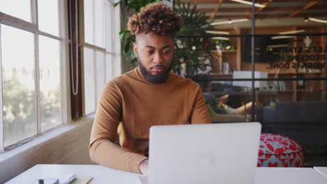 Millennial-black-male-blogger-using-laptop--at-a-desk-by-the-window-in-a-creative-office,-close-up