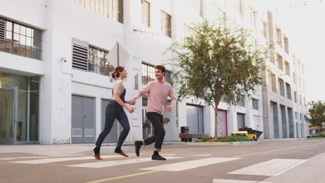 Millennial-hipster-couple-running-across-the-road-in-the-city-holding-hands,-full-length
