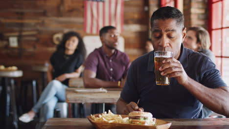 Mature-Man-Watching-Game-On-Screen-In-Sports-Bar-Eating-Burger-And-Fries-And-Drinking-Beer