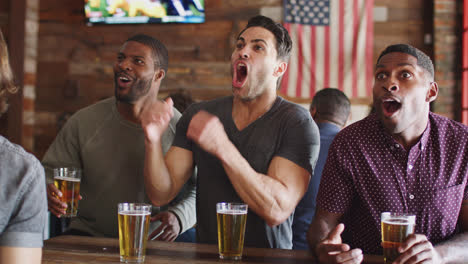 Group-Of-Male-Friends-Celebrating-Whilst-Watching-Game-On-Screen-In-Sports-Bar