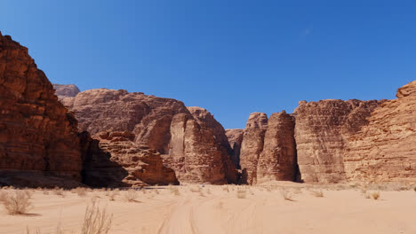 White-sand-below-river-valley-of-Wadi-Rum,-epic-cliffs-rise-against-blue-sky