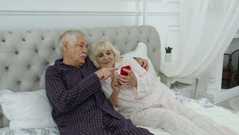 Senior-elderly-couple-wearing-pyjamas-lying-on-bed-looking-on-mobile-phone-laughing-and-having-fun