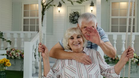 Senior-elderly-couple-in-front-yard-at-home.-Man-hugging-woman.-Happy-mature-family-waving-hands