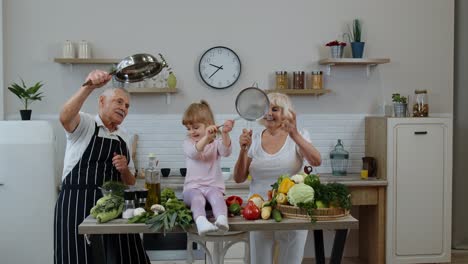 Senior-woman-and-man-with-grandchild-girl-making-a-funny-dance-with-strainer-and-vegetables-at-home
