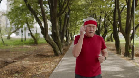 Male-senior-person-running-along-the-road-in-park.-Mature-runner-man-training,-listening-music