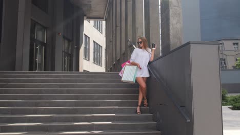 Girl-with-phone-staying-on-shopping-mall-stairs-with-colorful-shopping-bags-and-drinking-coffee