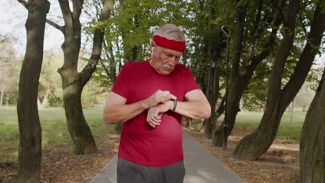 Anciano-Corriendo-En-El-Parque-Y-Usando-Reloj-Inteligente,-Rastreando-El-Resultado-Después-Del-Entrenamiento-Físico