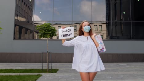 Girl-in-protective-mask-with-shopping-bags-showing-Black-Friday-inscription-during-coronavirus