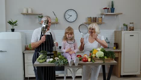 Senior-woman-and-man-with-grandchild-girl-making-a-funny-dance-with-strainer-and-vegetables-at-home