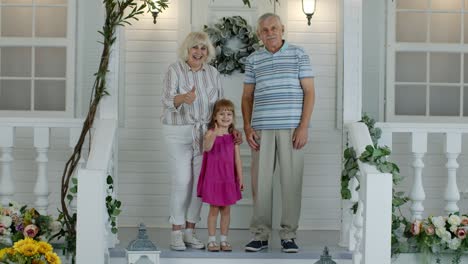 Senior-grandparens-couple-with-granddaughter-in-porch-at-home-waving-hand,-showing-thumbs-up