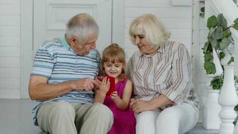 Senior-couple-sitting-with-granddaughter-and-using-mobile-phone-in-porch-at-home.-Online-shopping