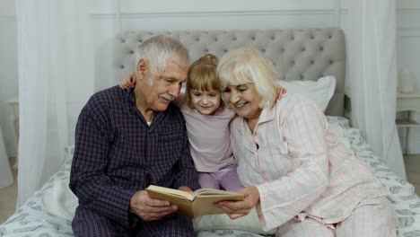 Süßes-Mädchen-Mit-älterer-Großmutter-Und-Großvater-Im-Ruhestand,-Die-Auf-Dem-Bett-Sitzen-Und-Im-Schlafzimmer-Ein-Buch-Lesen
