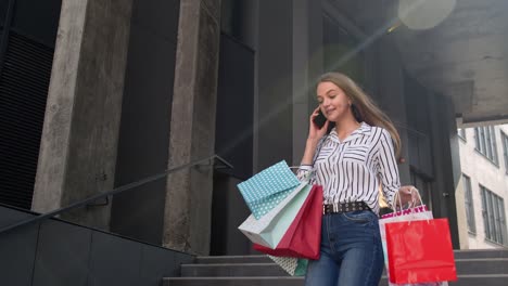 Girl-walking-from-shopping-mall-with-shopping-bags-and-talking-on-mobile-phone-about-purchases