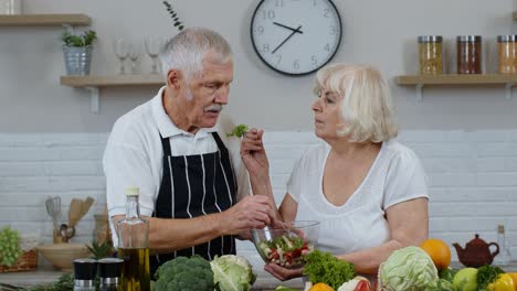 Senior-couple-in-kitchen.-Grandmother-and-grandfather-feeding-each-other-with-raw-vegetable-salad