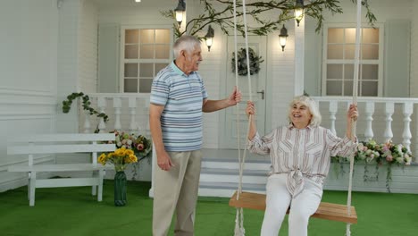 Senior-couple-together-in-front-yard-at-home.-Man-swinging-woman.-Happy-elderly-pensioners-family