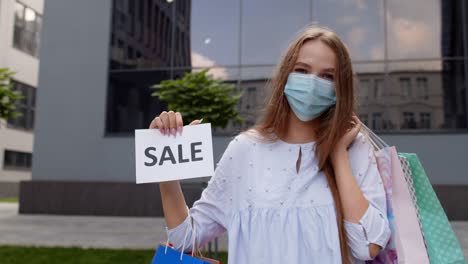 Girl-in-protective-mask-with-shopping-bags-showing-Sale-word-inscription-during-coronavirus-pandemic