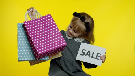 Cute-joyful-kid-girl-in-shirt-showing-Sale-word-advertisement-inscription-banner-and-shopping-bags