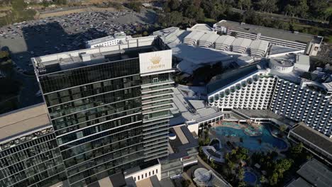 Luftaufnahme-Von-Oben-Nach-Unten-Des-Luxuriösen-Crown-Casino-Hotels-In-Perth-City,-Beleuchtung-Bei-Sonnenuntergang-Und-Parkende-Autos-Im-Hintergrund