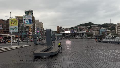 People-sit-and-walk-at-the-Keelung-Maritime-Plaza-waterfront-against-the-background-of-the-city-landmarks-and-iconic-Keelung-sign-in-Keelung-City,-Ren’ai-District,-Taiwan