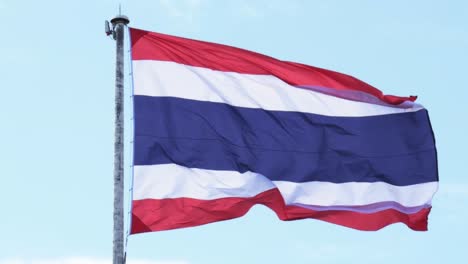Thailand-national-flag-hoisted-on-a-white-tall-steel-pole-blown-by-the-wind-creating-an-animated-wave-on-a-backdrop-of-a-clear-blue-sky-and-white-clouds