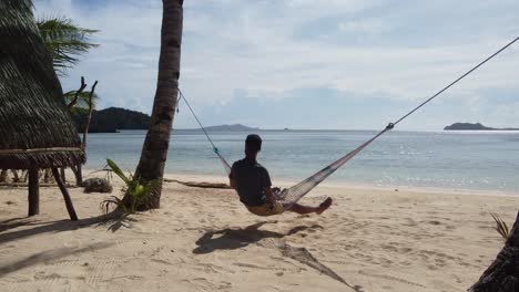 Young-adult-swing-on-tree-hammock-at-tropical-sunny-sand-beach-beside-bungalow