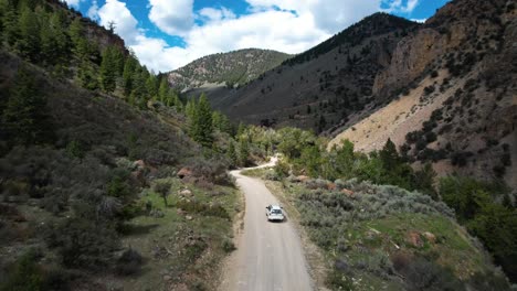 White-Car-drives-up-dirt-road-into-the-mountains-in-summertime-with-pine-trees