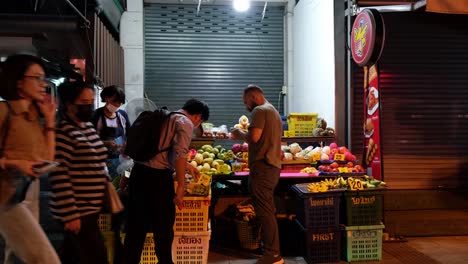 Dos-Personas-Ocupadas-Eligiendo-El-Tipo-Correcto-De-Frutas-Para-Comprar-En-Una-Acera-En-Bangkok-Mientras-Pasan-Los-Viajeros,-Bangkok,-Tailandia