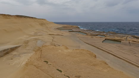 Luftaufnahme-über-Salzpflanzen-Malta-Oder-Salzverdunstungspfannen-An-Maltas-Felsiger-Küste-Mit-Trockener-Landschaft-Mit-Blick-Auf-Das-Meer-Und-Ruhigen-Wellen-An-Einem-Bewölkten-Tag