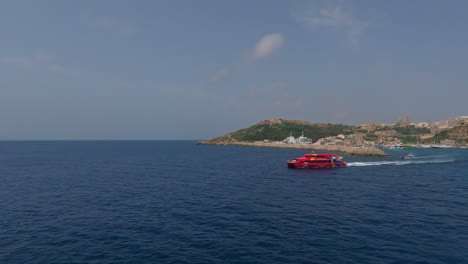 Luftaufnahme-über-Die-Wunderschöne-Küste-Von-Mgarr-Auf-Malta-Mit-Blick-Auf-Eine-Rote-Fähre,-Die-Touristen-Auf-Eine-Bootsfahrt-Durch-Das-Meer-Zum-Nächsten-Hafen-Und-Der-Insel-Im-Hintergrund-Bringt