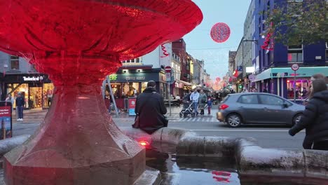 Street-with-red-lit-water-fountains-and-Christmas-decorations-in-Cork-city,-Ireland-with-traffic-and-people