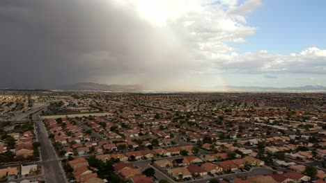 Aerial-footage-of-North-Las-Vegas-shows-rows-of-homes-under-stormy-skies,-with-a-distant-rainbow-and-mountains-contrasting-the-suburban-expanse