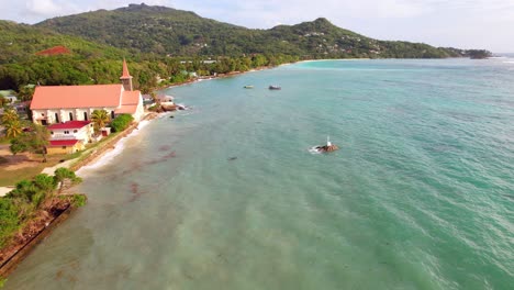 Drone-of-St-Joseph-church-near-the-shore-of-Anse-royale-beach,-cross-on-rock-in-the-centre-of-the-ocean,-Mahe,-Seychelles-30fps