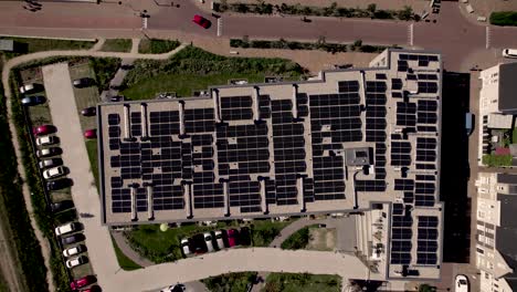 Top-down-aerial-view-of-apartment-complex-roof-full-of-solar-panels-with-cars-stationed-and-passing-by-on-the-street-sharing-the-road-with-bikers