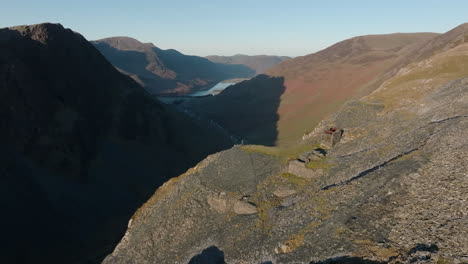 Flying-high-over-mountain-walker-stood-on-cliff-edge-at-dawn-over-the-Honister-Pass-with-Buttermere-in-the-distance